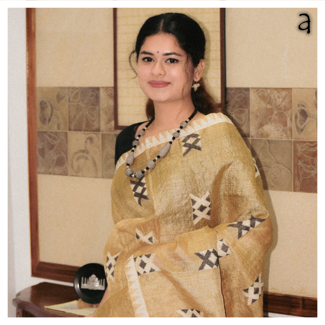 Woman in a yellow saree with black patterns sitting indoors.
